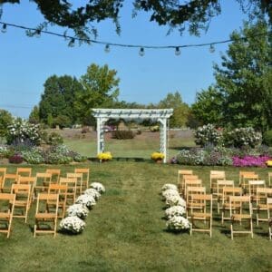 Outdoor Garden Venue setup with rows of wooden chairs facing a white pergola, surrounded by colorful flowers and strung lights above on a sunny day.