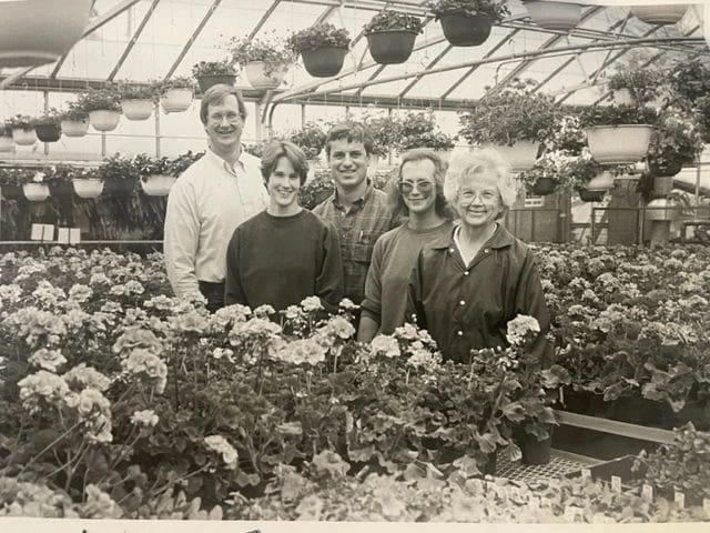 Five people smiling in a greenhouse surrounded by flowering plants and hanging baskets.