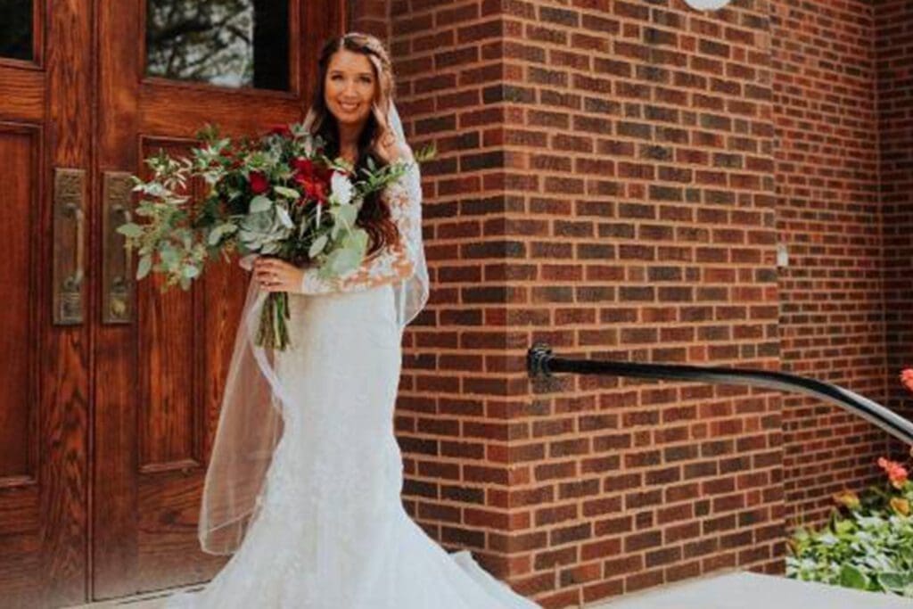 A bride in a white lace gown, smiling and holding a bouquet of red and white flowers, stands beside a brick building.