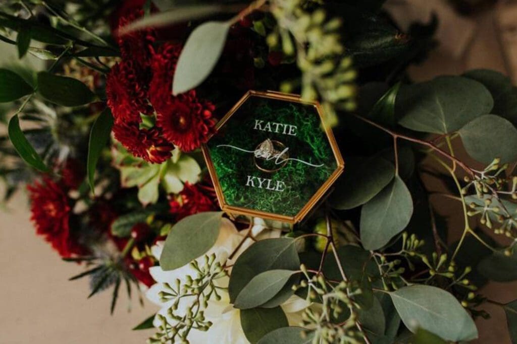 A hexagonal wedding ring box with engraved names "katie & kyle" surrounded by a floral arrangement with red flowers and green leaves.