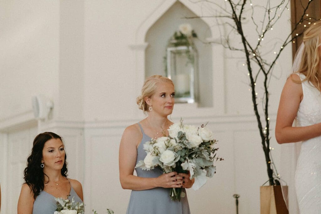 A bridesmaid in a pale blue dress, holding a bouquet, looks towards the bride inside a church decorated with fairy lights. another bridesmaid is partially visible.