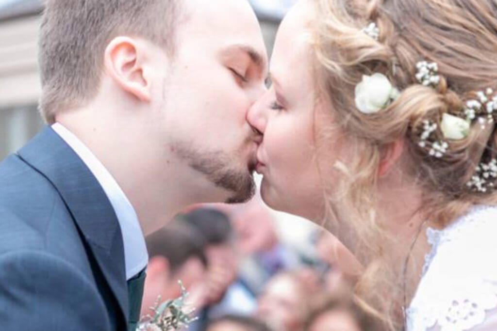 A bride and groom kissing, with the bride wearing a white dress adorned with floral elements in her hair and the groom in a gray suit.