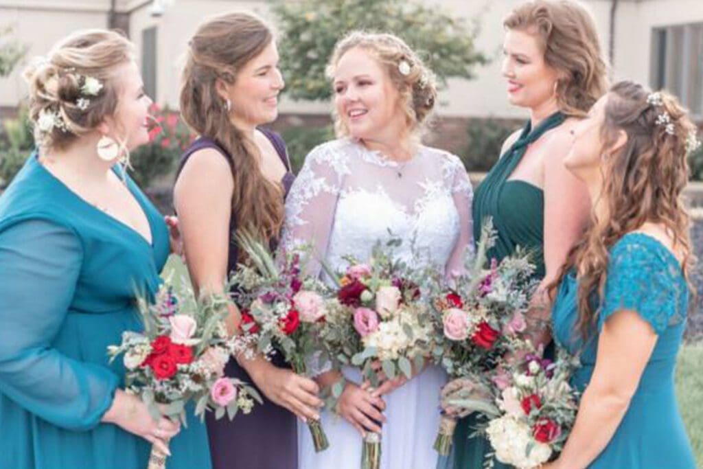 Five women in formal dresses with floral bouquets and hairpieces smiling and chatting at an outdoor wedding event.