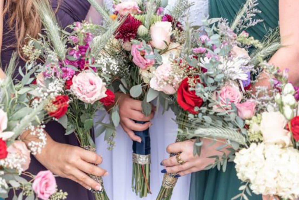 Close-up of three women holding colorful bridal bouquets, featuring pink and white roses, with a focus on their hands and the floral arrangements.