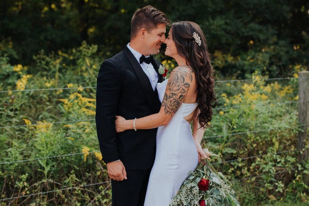 A bride and groom smiling at each other in a field, the bride holding a bouquet and showcasing a tattooed arm.