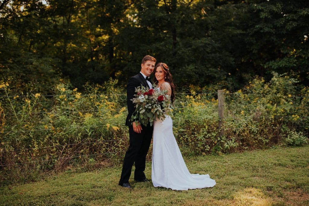A newlywed couple posing outdoors, the man in a black suit and the woman in a white dress, holding a bouquet, with trees and bushes in the background.