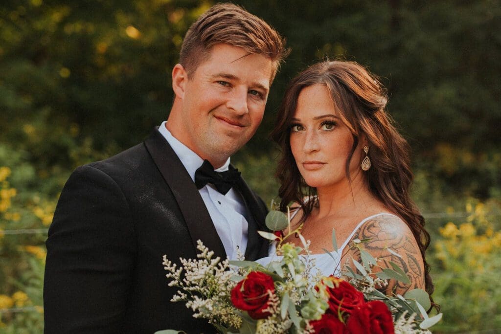 A bride and groom in formal attire, holding a bouquet, with a natural backdrop during golden hour.