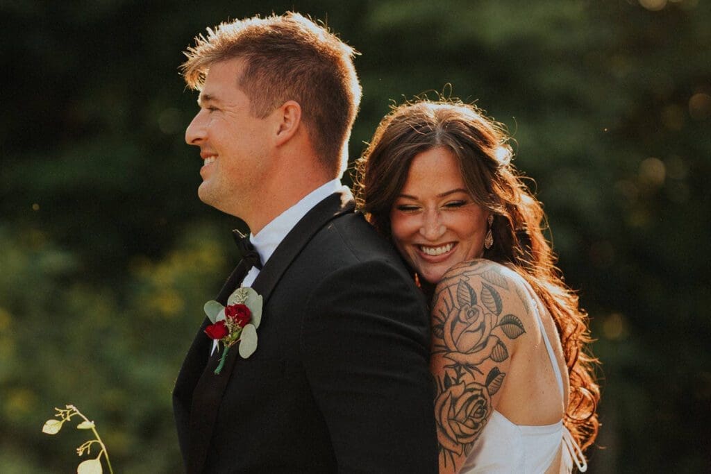 A bride with a tattoo on her arm hugs a groom from behind, both smiling in a sunlit outdoor setting.