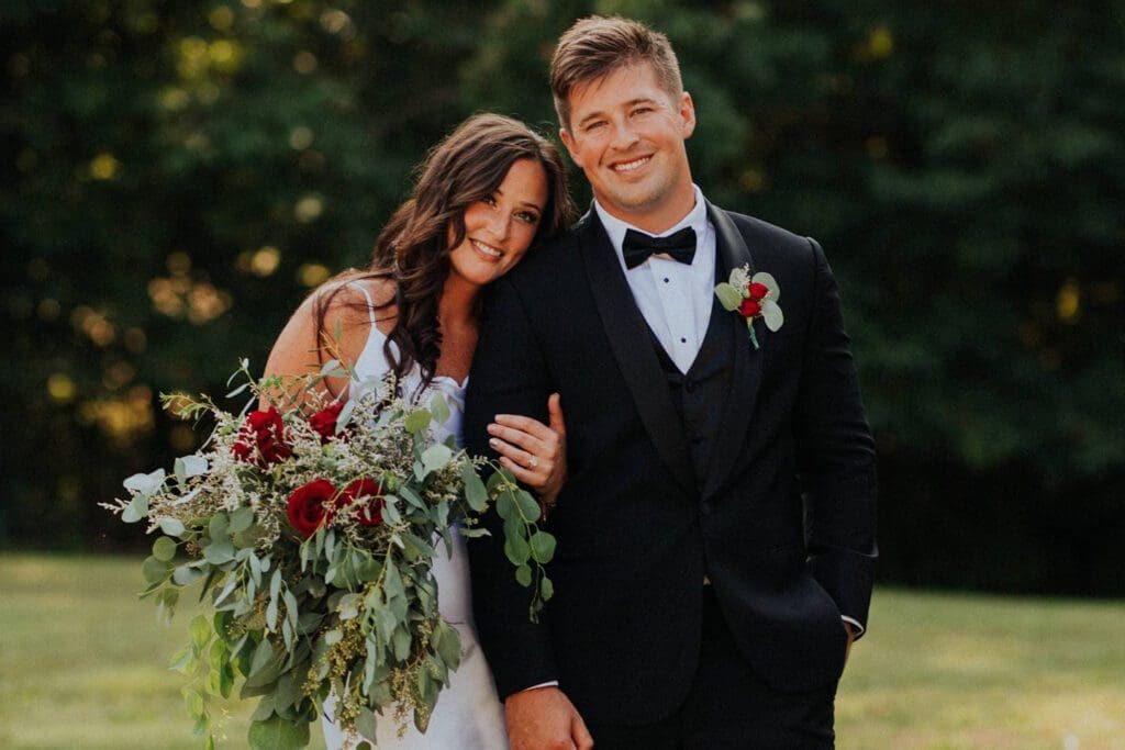 A bride and groom smiling and posing together at their wedding, the bride holding a large bouquet, in an outdoor setting.