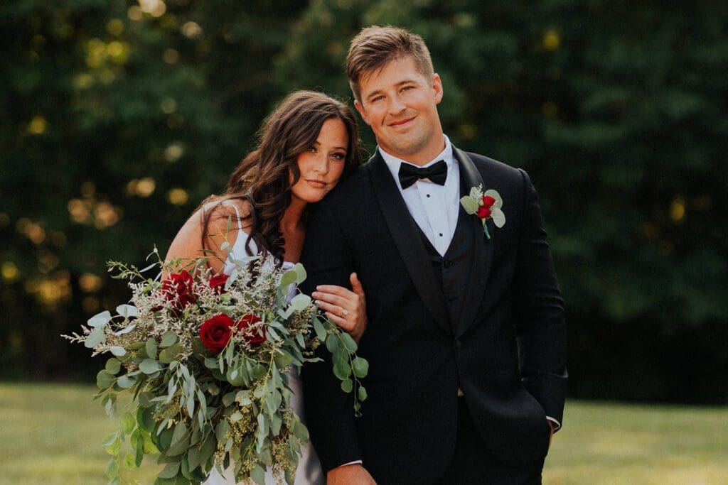 A bride and groom smiling outdoors, the bride holding a large bouquet.