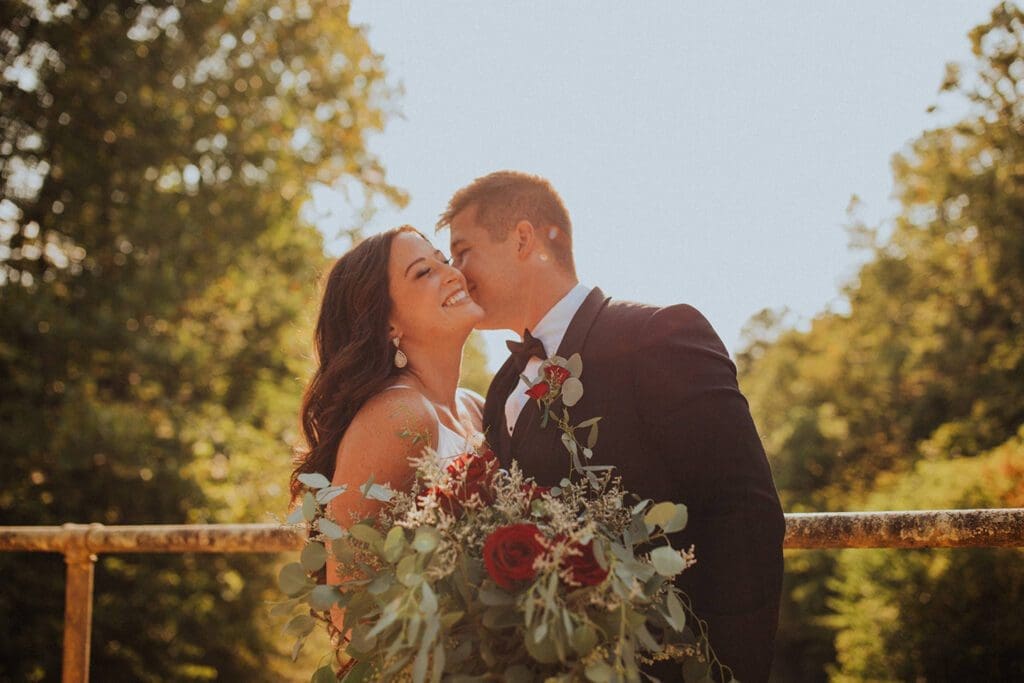 A bride and groom share a kiss on a sunny day, the bride holding a bouquet of red flowers, surrounded by lush greenery.