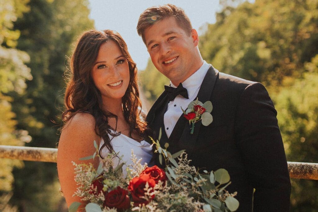 A bride and groom smiling outdoors, the bride holding a bouquet with red flowers, both dressed in wedding attire.