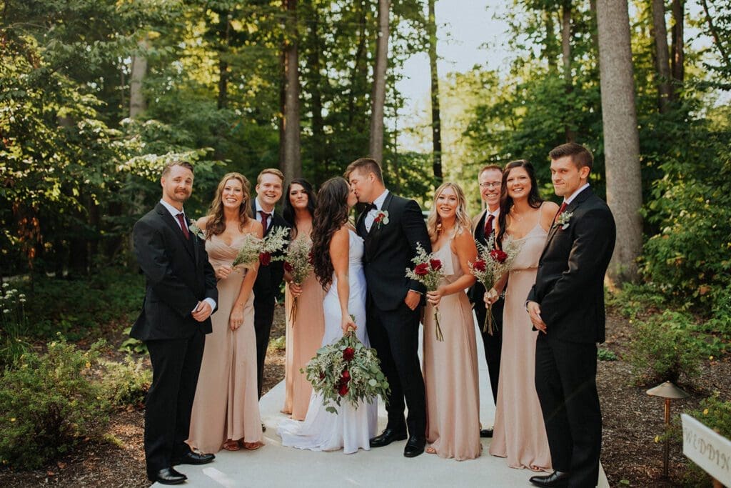 A wedding party standing outdoors, featuring a bride and groom at the center, flanked by bridesmaids in beige dresses and groomsmen in suits.