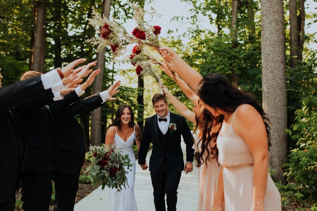 Newlyweds walk down a path while friends toss flowers overhead; the bride and groom look happy and festive.