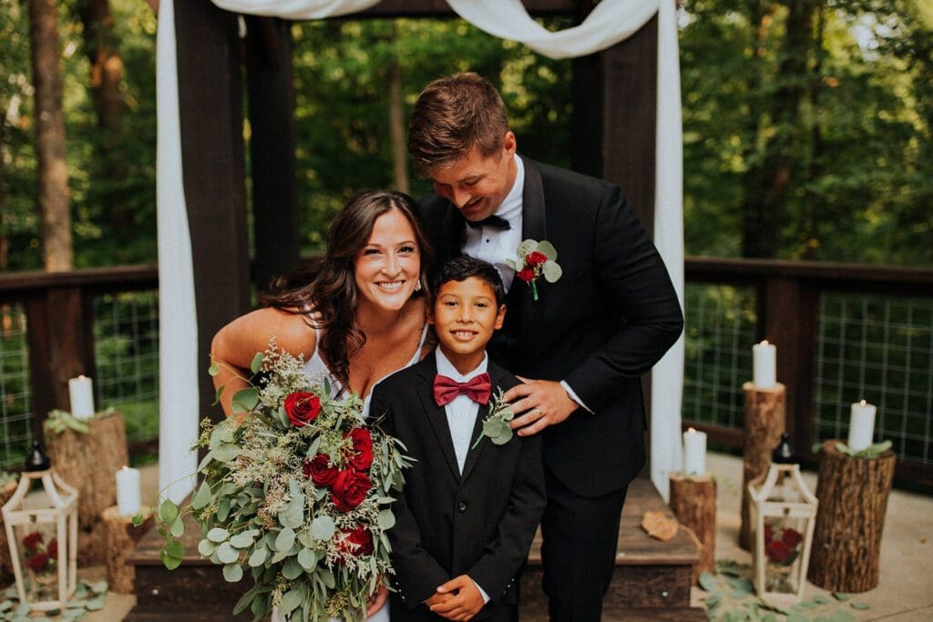 A bride and groom smile alongside a young boy in formal attire, posing under an outdoor wedding arch decorated with candles.