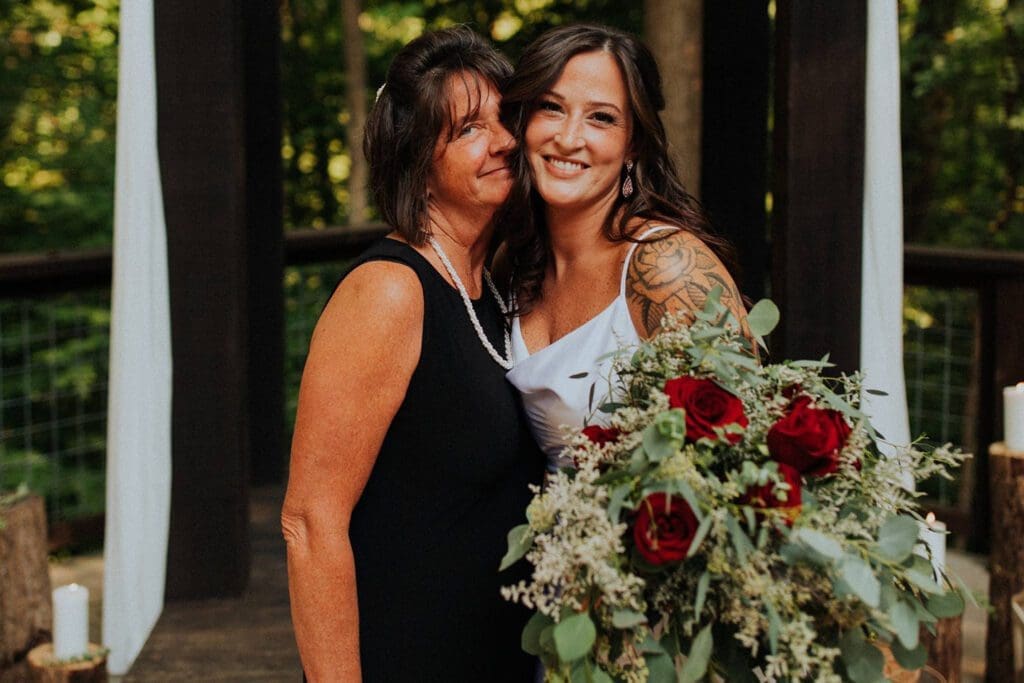Two smiling women, one younger with a tattoo and holding a bouquet of red roses, and the older one in a black dress, standing together in a wooded area.