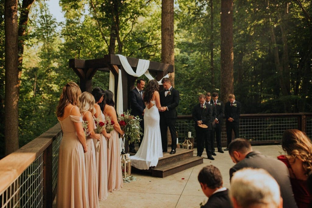 A wedding ceremony in a forest setting, with a couple exchanging vows at an altar, surrounded by guests and bridesmaids in peach dresses.
