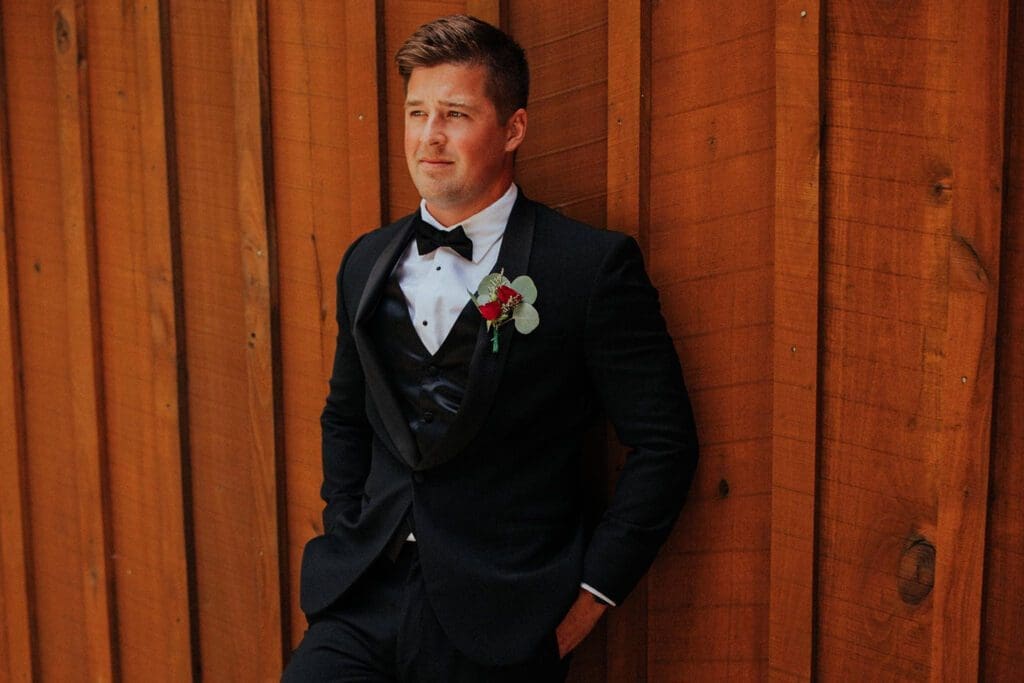Man in a black tuxedo and bow tie leaning against a wooden wall, looking to the side with a thoughtful expression. he wears a boutonniere with a red flower.