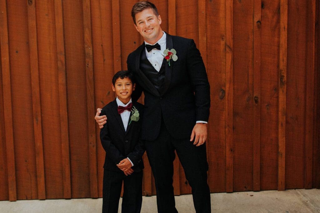 A man and a young boy in tuxedos, smiling, stand in front of a wooden fence.