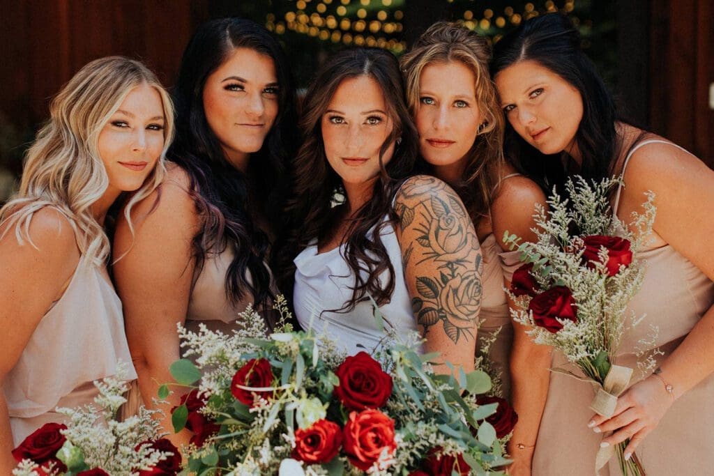 Five women posing closely together, holding a floral arrangement with red roses, in an outdoor setting. they wear matching beige dresses and the bride stands at the center in white.