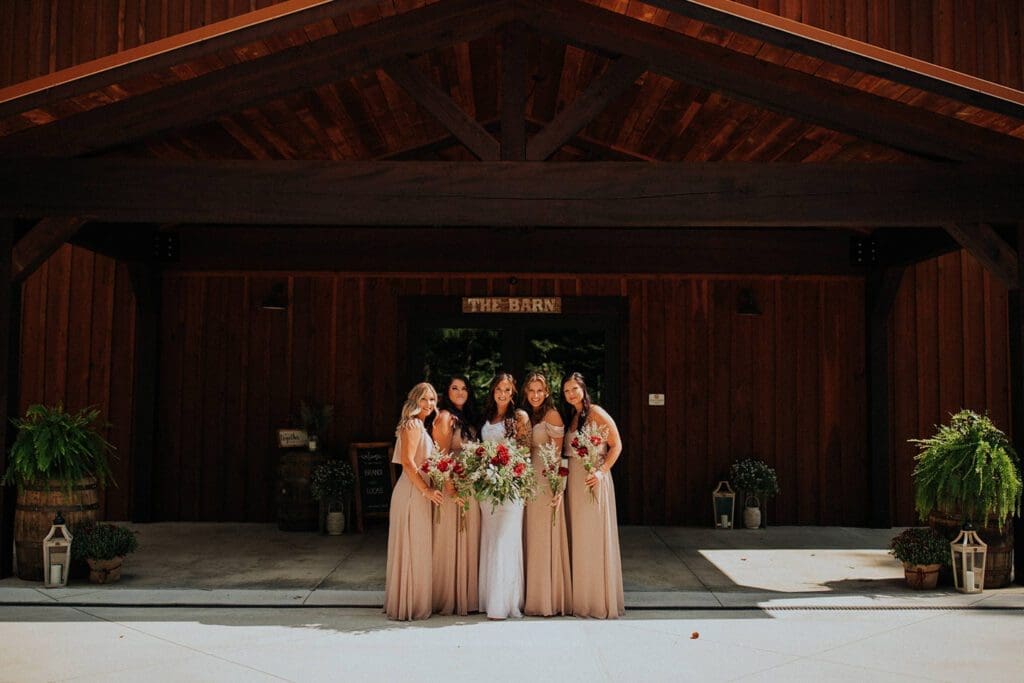 Five women in matching beige dresses holding bouquets, standing in front of a barn entrance labeled "the barn.