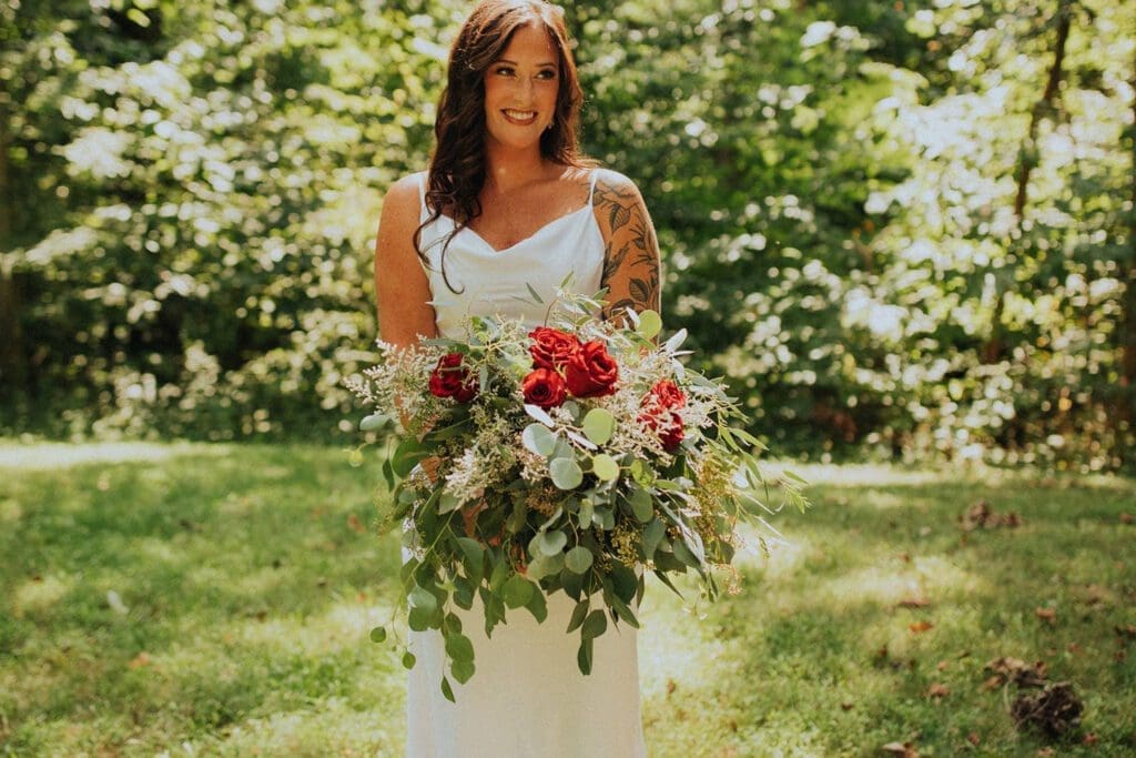 A smiling woman in a white dress holding a large bouquet with red flowers and green foliage, standing in a sunlit park.