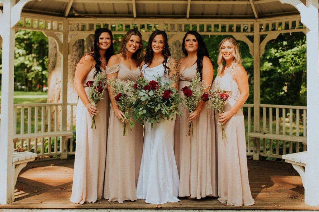 Five women in coordinated dresses, holding bouquets, pose together in a gazebo surrounded by trees.