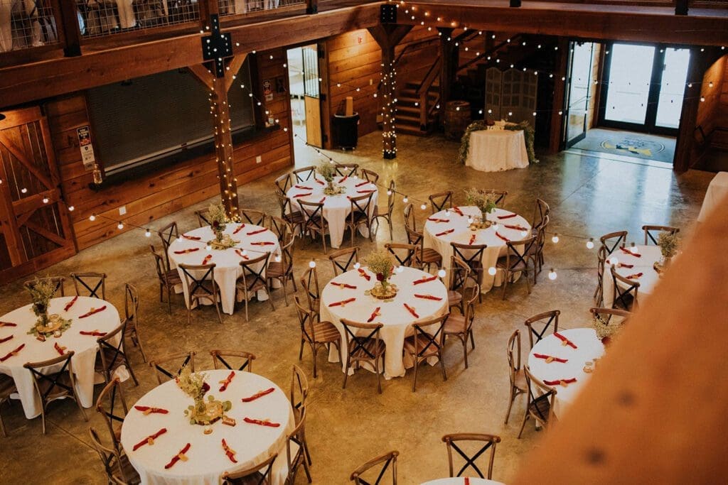 Elegantly arranged wedding reception inside a rustic wooden barn, featuring round tables with white linens and red napkins.