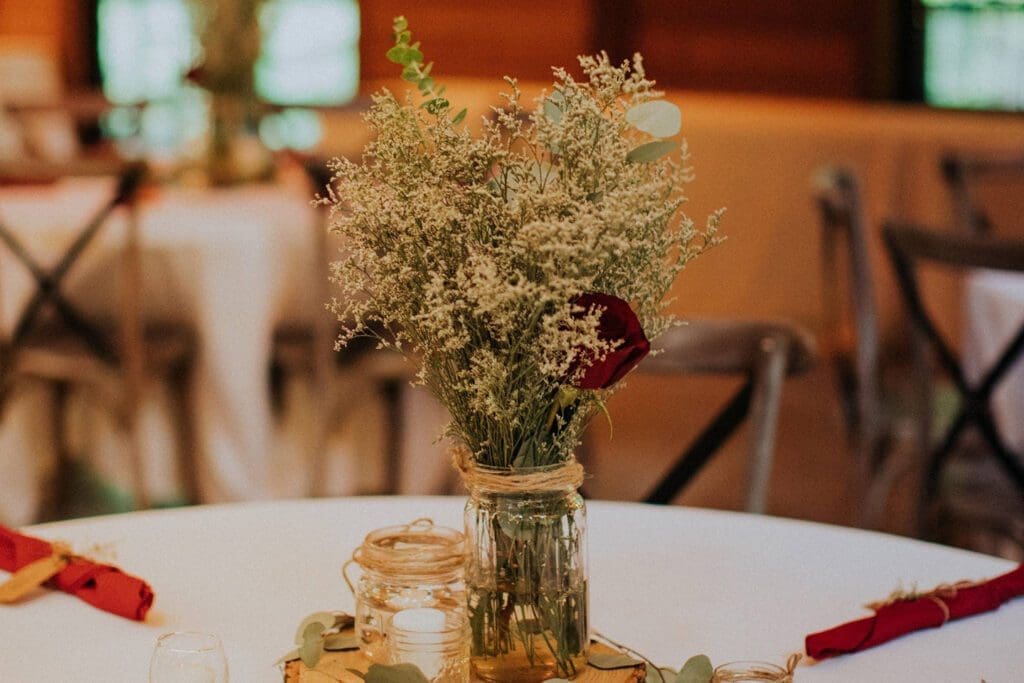 A rustic table setting with a mason jar floral arrangement, surrounded by votive candles and scattered leaves, in a warmly lit banquet hall.