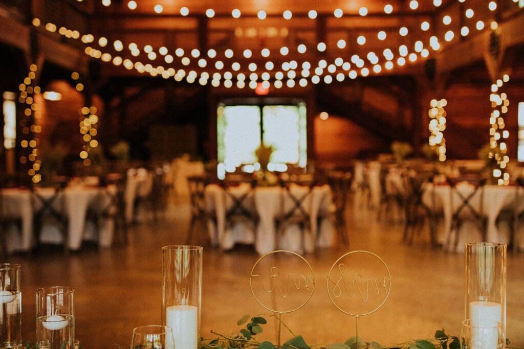 Rustic wedding reception hall decorated with string lights and tables set for dining, featuring "bride" and "groom" signs in the foreground.