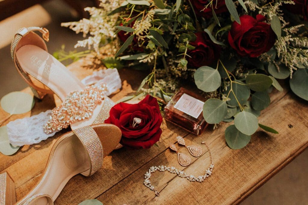A stylish bridal arrangement with beige shoes, a red rose bouquet, perfume, and jewelry on a wooden table.