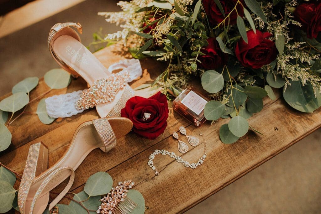 Elegant bridal accessories laid out on a wooden surface, including high-heeled shoes, a bouquet of red roses, earrings, and a necklace.