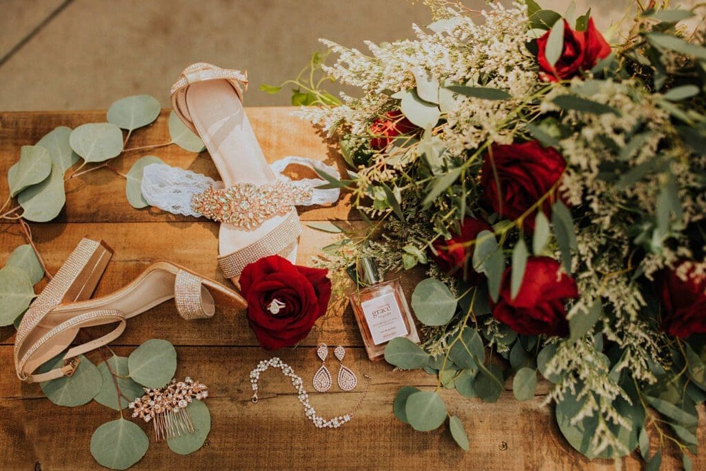 A wedding setup with a pair of high-heel sandals, red roses, green foliage, jewelry, and a perfume bottle on a wooden table.