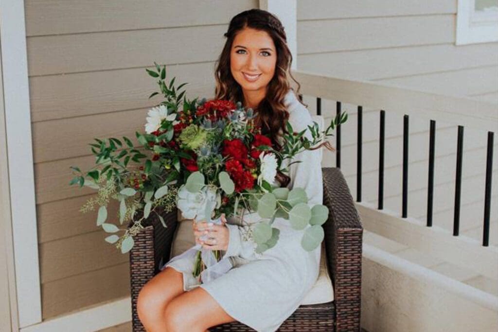 A woman in a white dress smiling and holding a large bouquet of flowers while seated on a porch bench.