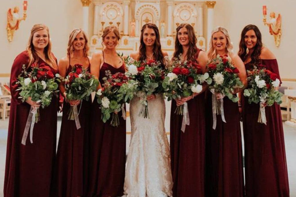A bride in a white dress stands with six bridesmaids in dark red dresses, all holding large bouquets, inside a church.