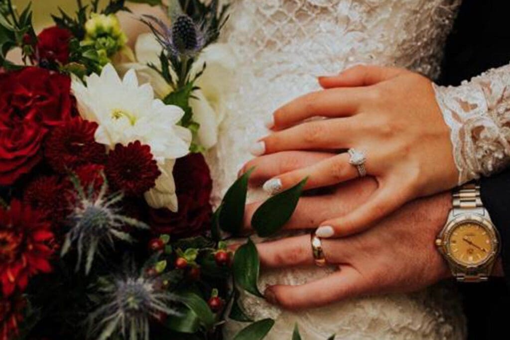 A close-up of a newlywed couple's hands over a bouquet, showcasing their wedding rings and elegant attire, with a focus on the delicate details of the flowers and jewelry.