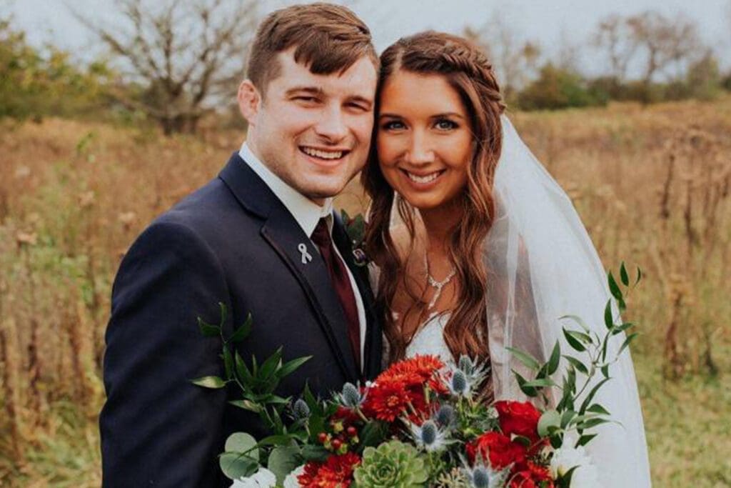 A newlywed couple smiling outdoors with the bride in a veil holding a bouquet of red and green flowers, and the groom in a navy suit with a boutonniere.