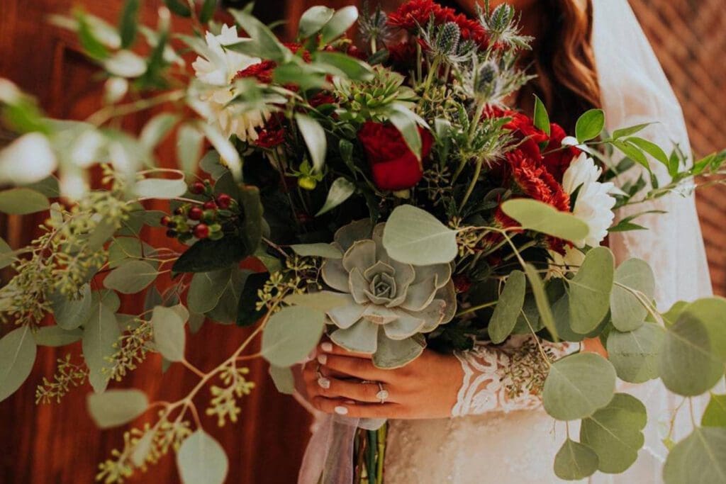 Bride holding a vibrant bouquet with red roses and green succulents, framed by her lace sleeve.