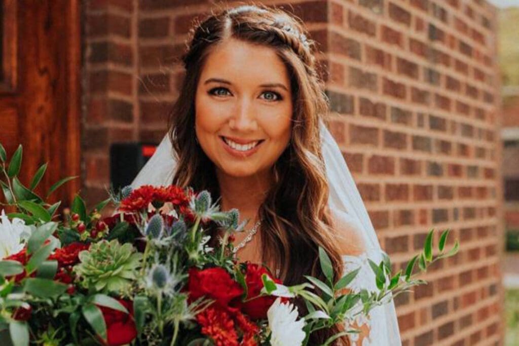 A smiling bride holding a bouquet of red and white flowers stands in front of a brick wall on her wedding day.